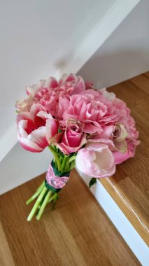 Yumuşak pembe karanfillerden ve lalelerden oluşan bir buket ahşap bir yüzey üzerinde duruyor. Sapları dekoratif pembe ve küçük bir düğmeyle süslenmiş siyah kumaşla sarılmış. Arkaplan beyaz, canlı pembe renklere zıtlık sağlıyor