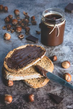 Kavanozda ev yapımı fındıklı çikolatalı tereyağı ve bir parça tam buğday ekmeğine, fındıklı fındıklı çikolataya sürülmüş siyah taş arka planda..