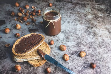 Kavanozda ev yapımı fındıklı çikolatalı tereyağı ve bir parça tam buğday ekmeğine, fındıklı fındıklı çikolataya sürülmüş siyah taş arka planda..