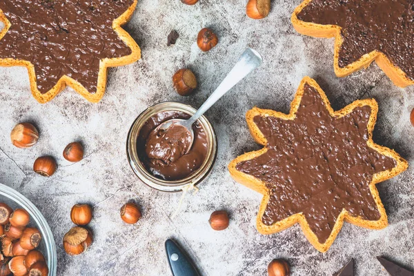 Kavanozda ev yapımı fındıklı çikolatalı tereyağı ve yıldız şeklinde pandoro panettone, fındık, koyu çikolata parçalarına yayılmış siyah taş arka planda. Üst Manzara. Noel yemeği konsepti.