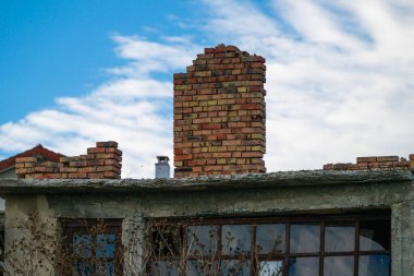 A weathered rooftop with brick chimneys rises above broken windows and dry vegetation, capturing the quiet decay of an abandoned building under a partly cloudy sky.