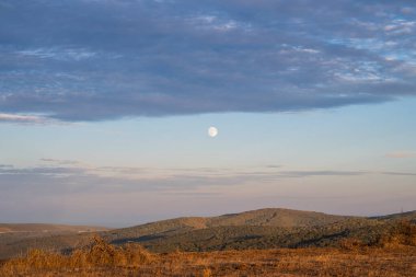 A pale moon hangs quietly above rolling hills and dry grass in the mountains in Bulgaria
