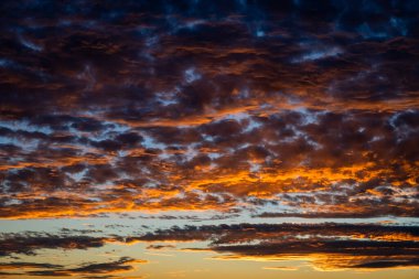 Kızgın bulutlar, gün batımında dramatik bir gökyüzünde kıvrılır, derin maviler, morlar, portakallar ve altın sarılarını harmanlayarak nefes kesici bir görüntüde.