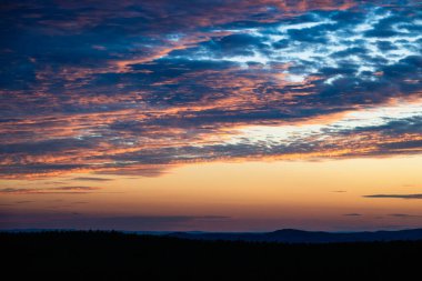 Kızgın bulutlar, gün batımında dramatik bir gökyüzünde kıvrılır, derin maviler, morlar, portakallar ve altın sarılarını harmanlayarak nefes kesici bir görüntüde.