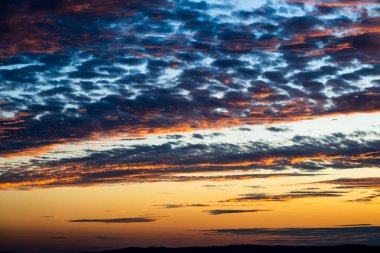 Kızgın bulutlar, gün batımında dramatik bir gökyüzünde kıvrılır, derin maviler, morlar, portakallar ve altın sarılarını harmanlayarak nefes kesici bir görüntüde.