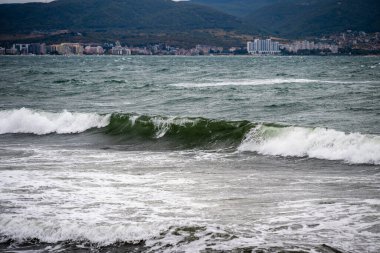 Dalgalar fırtınalı gökyüzünün altında kayalık bir sahile çarpıyor ve Bulgaristan 'daki tepeler ve denizler arasında uzak bir şehir manzarası var.