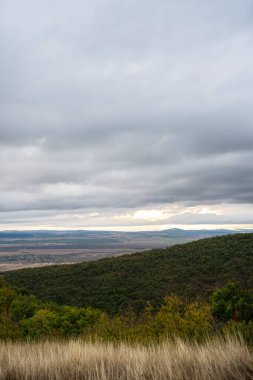 Güneş ışığı, yuvarlanan tepeler ve kuru otlar üzerinde yoğun gri bulutları yararak kasvetli bir sonbahar gökyüzünün altında geniş bir ovayı ortaya çıkarıyor..