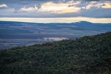 Ormanlık tepelerin altındaki güneşli bir vadide sis sürükleniyor ve dramatik, bulutlu bir gökyüzü.