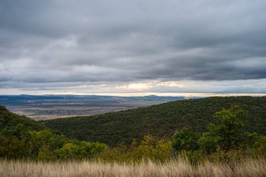 Ormanlık tepelerin altındaki güneşli bir vadide sis sürükleniyor ve dramatik, bulutlu bir gökyüzü.
