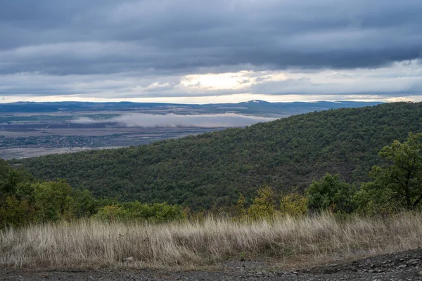 Ormanlık tepelerin altındaki güneşli bir vadide sis sürükleniyor ve dramatik, bulutlu bir gökyüzü.