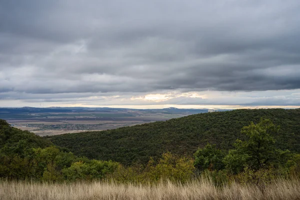 Ormanlık tepelerin altındaki güneşli bir vadide sis sürükleniyor ve dramatik, bulutlu bir gökyüzü.