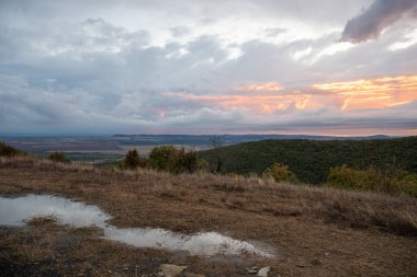 Güneş ışığı, ormanlık bir tepenin üzerindeki fırtınalı bulutları delip geçiyor. Su birikintisi yollara ve uzak ovalara altın ışık saçıyor..