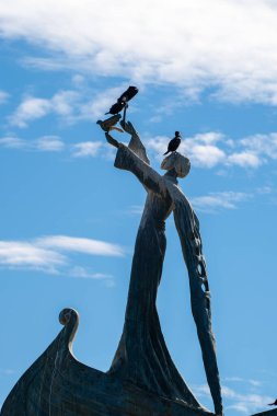  Kuşları tutan cüppeli bir heykelin bronz heykeli, dağınık bulutlu canlı mavi gökyüzünün altında stilize edilmiş bir teknenin pruvasında duruyor..
