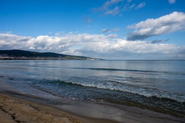 Yumuşak dalgalar bulutlu bir gökyüzünün altında kumlu bir sahille buluşuyor. Uzak tepeler ve ufku kaplayan bir sahil kasabası var..