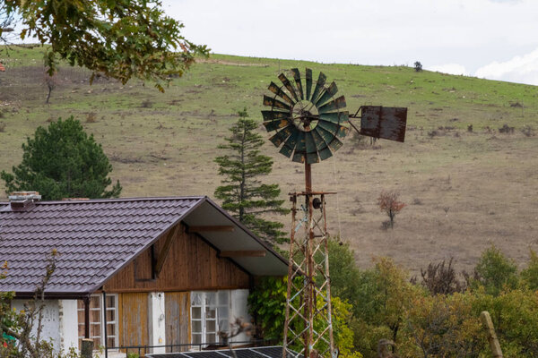 A weathered windmill and rustic house nestle into a grassy hillside, evoking the quiet charm of rural life.