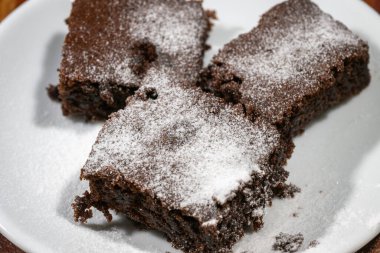 Üç zengin çikolatalı brownie pudra şekeri ile tozlanmış. Kaba ahşap bir yüzeyde cezbedici bir şekilde dinleniyor..
