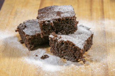 Üç zengin çikolatalı brownie pudra şekeri ile tozlanmış. Kaba ahşap bir yüzeyde cezbedici bir şekilde dinleniyor..
