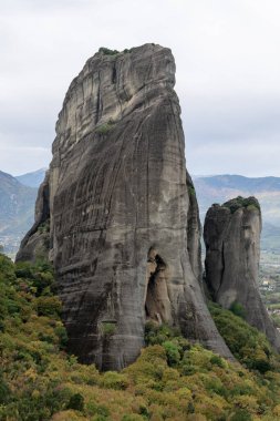 Meteora 'da parçalı bulutlu bir gökyüzünün altında, dağlık bir arazide, bereketli bir ormandan uzun, yıpranmış kaya sütunları yükselir.