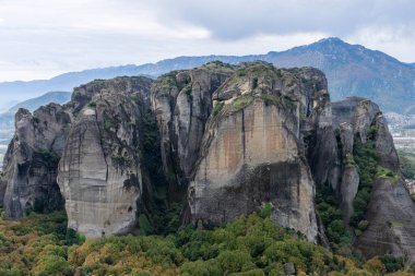 Dağlık bir araziden yükselen yüksek bir kaya oluşumu engebeli uçurumları bulutlu bir gökyüzünün altındaki dağınık yeşilliklerle yumuşadı..