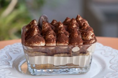 Bir cam tabak tiramisu sergileniyor kremalı katmanlar, kakao tozlu mascarpone, ve beyaz tabakta çikolatalı garnitür..