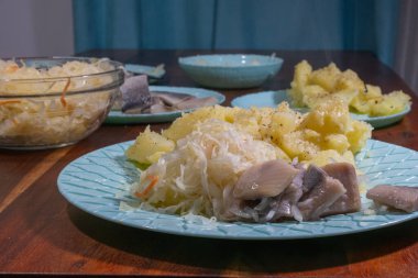 Bir tabak haşlanmış patates, lahana turşusu ve turşulu ringa balığı açık mavi bir tabakta servis edilir..