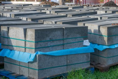 Trapezoidal beton kaldırım taşları paletlerin üzerinde duruyor, bir inşaat alanındaki açık hava deposuna bağlanmış ve bağlanmış..