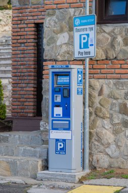 İki dilli bir park ödeme makinesi parçalı bulutlu bir binanın altında taş tuğlaların yanında duruyor.