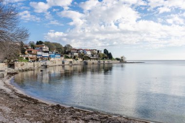 Gökyüzünü yansıtan sakin bir denizin sakin manzarası, parlak, bulutlu bir gökyüzünün altında küçük kumlu bir sahil ve kıyı evleri barındıran bir kıyı şeridi..