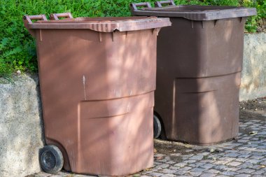 İki kahverengi çöp tenekesi beton duvarın ve yeşilliğin yanında asfalt üzerinde duruyor..