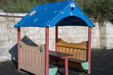 Boş, renkli, çocuk boyunda bir ahşap ve mavi çatılı plastik piknik sığınağı taştan bir duvara karşı park alanında duruyor..