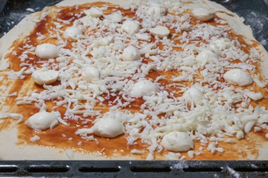 Bir pizza kabuğu kırmızı domates sosuyla kaplanır ve üzerinde büyük miktarda dilimlenmiş ve küçük mozzarella topları bulunur. Koyu bir fırın tepsisinde hazırlanır..