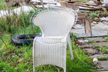 Beyaz, eski hasır bir sandalye, çimenli bir alanda dağınık enkaz arasında oturur, konfor ve ihmal arasındaki zıtlığı çağrıştırır.