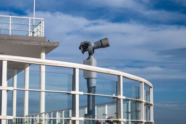 Bir monte edilmiş teleskop, açık bir gökyüzünün altındaki manzaralı bir güvertede eğimli bir korkuluğun arkasında duruyor.