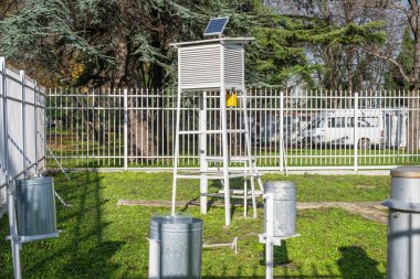 Stevenson ekranı ve yağmur göstergeleri olan bir meteoroloji istasyonu bulutlu gökyüzünün altında çimenli bir çitli alanda duruyor..