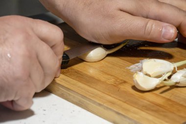 Bir insan mutfak bıçağı kullanarak tahta kesme tahtasının üzerinde taze sarımsak karanfilleri doğruyor..