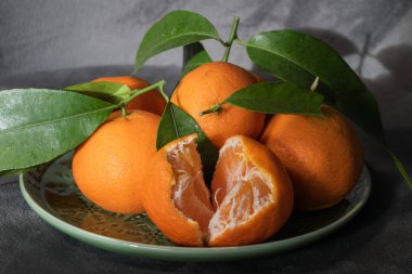Beş taze portakal, biri kısmen soyulmuş, seramik bir tabakta, parlak yeşil yapraklar ve sıcak ışık altında dinleniyor..
