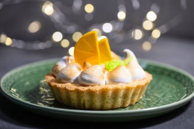 Kızarmış tepeleri ve turunçgilleri olan limonlu turta yeşil bir tabakta şenlikli ışıkların altında duruyor..