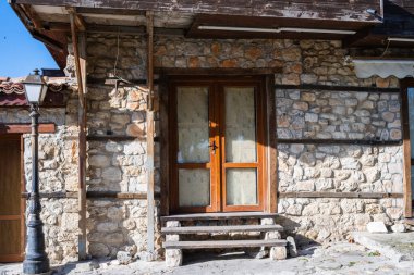 Ahşap çerçeveli cam kapılı taştan bir binaya güneşli bir çevrede kütük destekli basamaklardan ulaşılabilir..