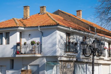 Kırmızı kiremitli çatısı olan beyaz bir binada, klasik bir sokak lambasıyla çerçevelenmiş, çiçekli ve bacalı balkonlar yer alıyor..