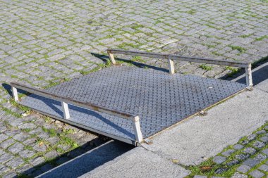 Elmas kaplama yüzeyi ve yan korkulukları olan küçük, taşınabilir metal bir rampa taştan bir kaldırımda duruyor..