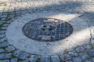 Şebeke desenli yuvarlak metal bir rögar kapağı eski kaldırım taşlarıyla çevrili beton bir halkanın içine yerleştirilir..