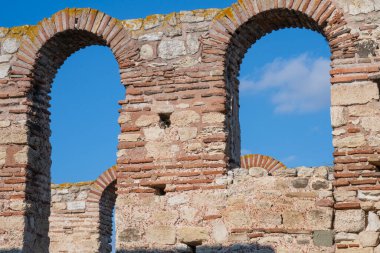  Yıpranmış bir taş ve tuğla duvardaki kemerli açıklıklar, temiz bir gökyüzünün altında antik zanaatkarlığı ortaya koyuyor..