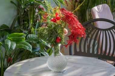 Kırmızı ve yeşil çiçekli şenlikli bir vazo, güneşli bahçedeki açık bir sandalyenin yanındaki beyaz mermer bir masanın üzerine yerleştirilir..