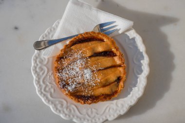 Pudra şekerli küçük, yuvarlak bir armut turtası beyaz bir peçetede duran çatalla süslü beyaz bir tabakta sunuluyor..
