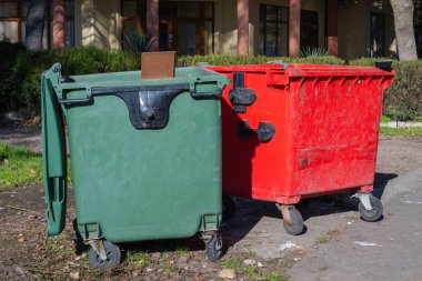 Biri yeşil diğeri kırmızı olmak üzere iki büyük endüstriyel atık kutusu. Arka planda ağaçlar ve bir bina bulunan asfalt bir alana yerleştirilmiş..