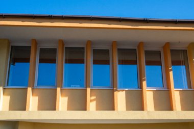 Açık mavi gökyüzünün altında dikey sarı-turuncu sütunlarla ayrılmış bir dizi dikdörtgen pencereli binanın ön cephesi.