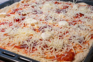 Çiğ, pişmemiş, üzerinde domates sosu, salam ve bol miktarda dilimlenmiş ve parçalanmış peynirle kaplı, siyah bir fırın tepsisindeki dikdörtgen pizza manzarası..