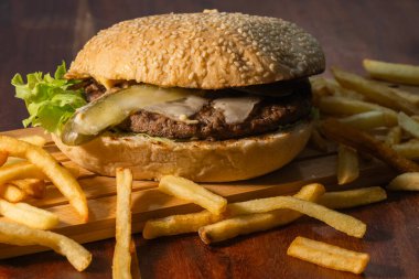 İçinde susamlı çörek, etli börek, turşu ve marul olan bir hamburger. Tahta bir tahtanın üzerinde dinleniyor. Etrafı koyu bir masada patates kızartmalarıyla çevrili..