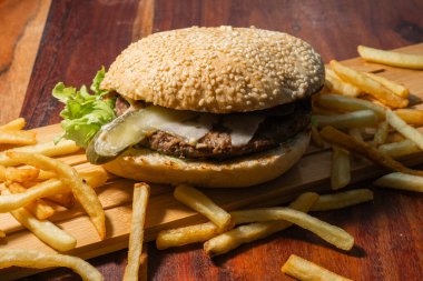 İçinde susamlı çörek, etli börek, turşu ve marul olan bir hamburger. Tahta bir tahtanın üzerinde dinleniyor. Etrafı koyu bir masada patates kızartmalarıyla çevrili..