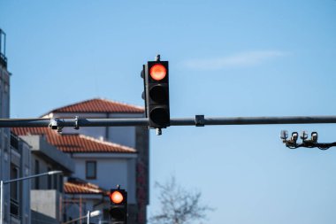 açık mavi gökyüzüne karşı metal bir kolda asılı duran bir trafik ışığı, arka planda fayanslı bir binanın çatısını gösteren parlak kırmızı bir sinyal verir..